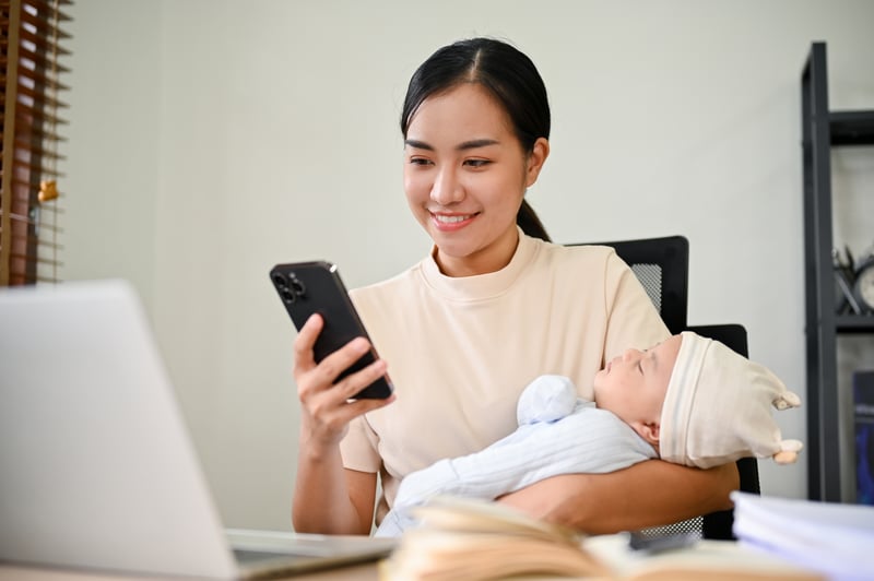 asian-mother-working-from-home-talking-phone-holding-her-baby-with-her-arms